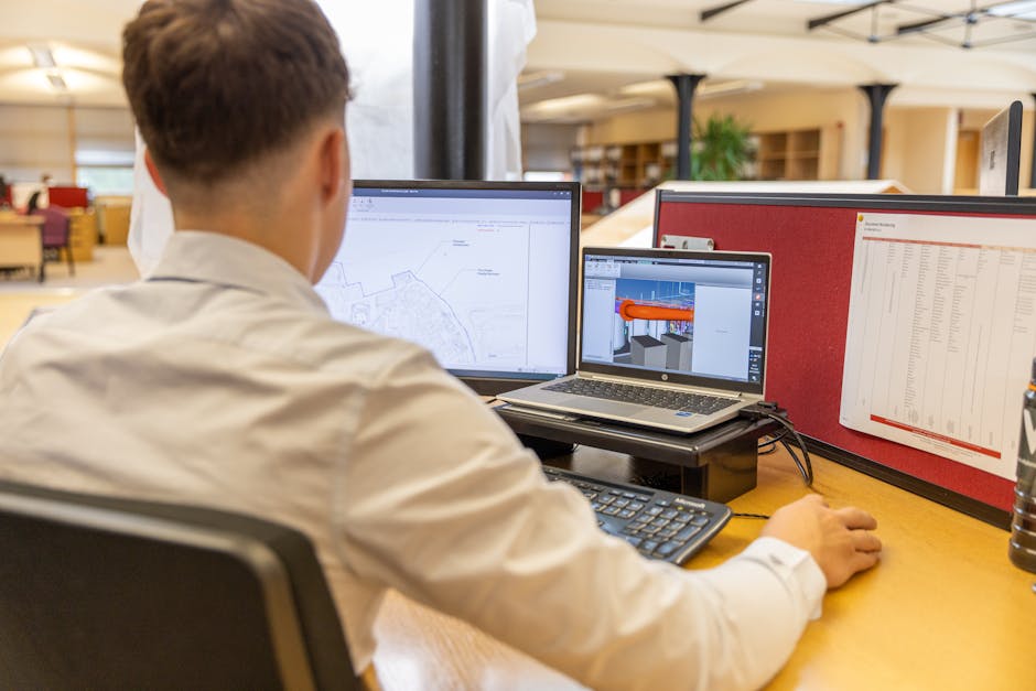 Back view of engineer focused on CAD design at dual monitors in bright office setting.