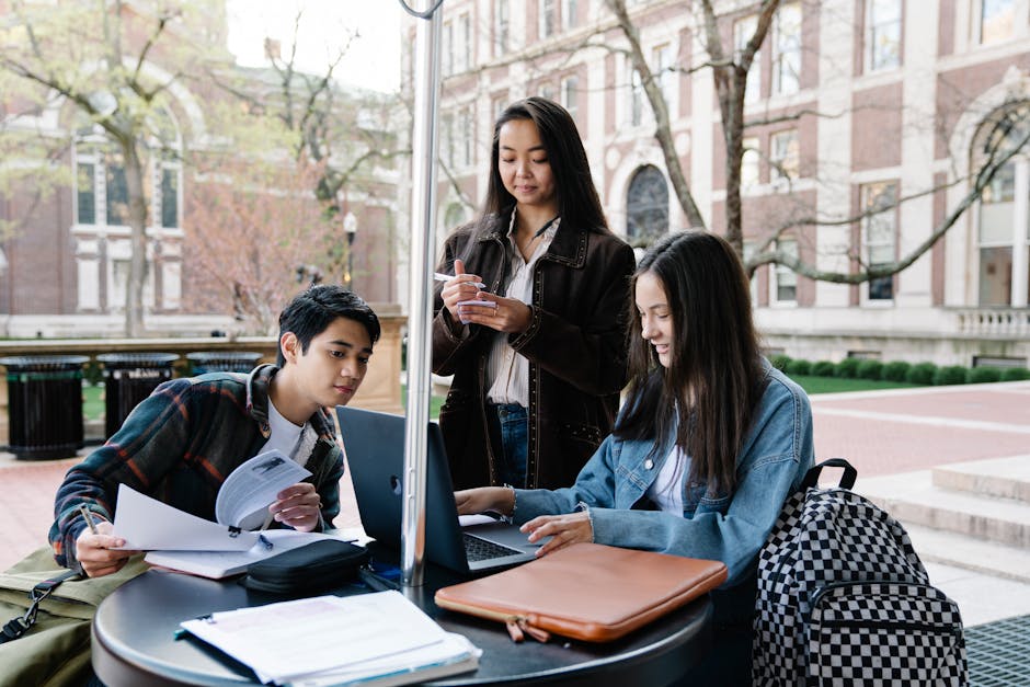 Students collaborate on a project outside a university building, engaged with their devices and materials.